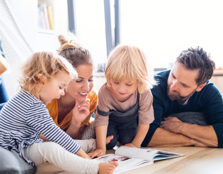 Front view of young family with two small children indoors in bedroom reading a book.